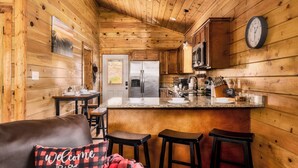 Una cocina acogedora de estilo cabaña con paredes de madera, un refrigerador de acero inoxidable, un microondas y un reloj en la pared. Hay una zona de bar con taburetes y un letrero de bienvenida.