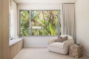 A room with a large window, a cushioned bench, a single chair, and a small side table.