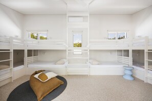 Habitación con literas, dos ventanas, una alfombra pequeña y un taburete azul.