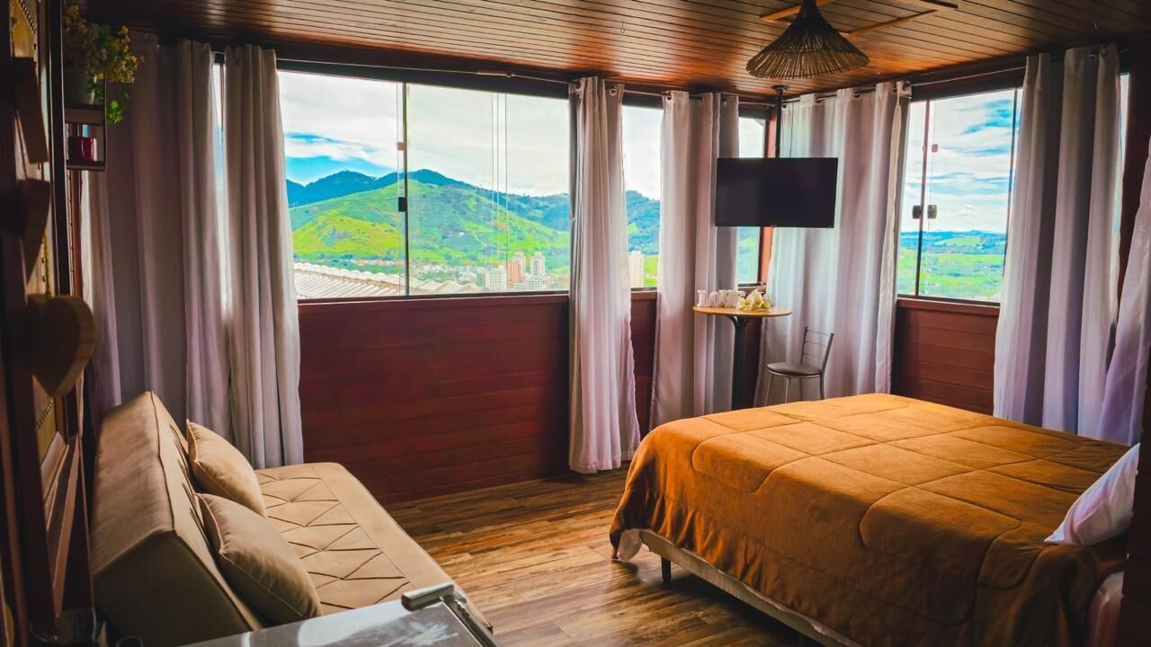 A room with a bed, a sofa, a TV, and a window with a view of green hills and buildings.