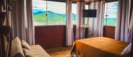 A room with a bed, a sofa, a TV, and a window with a view of green hills and buildings.