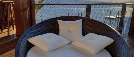 A wicker lounge chair with cushions on a wooden deck overlooking a mountainous landscape.
