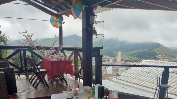An outdoor dining area with a table set for two, surrounded by wooden chairs, overlooking a cityscape with hills in the distance. - Chalezinho Pousada  (Santa Rita do Sapucaí)
