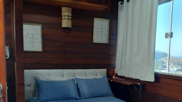Una habitación tipo cabaña de madera, con una cama, una ventana con cortinas y dos cuadros en la pared.