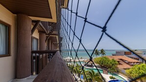 Balcón con vista a una piscina y una playa, con una barandilla de seguridad de malla y un pasamanos de madera.
