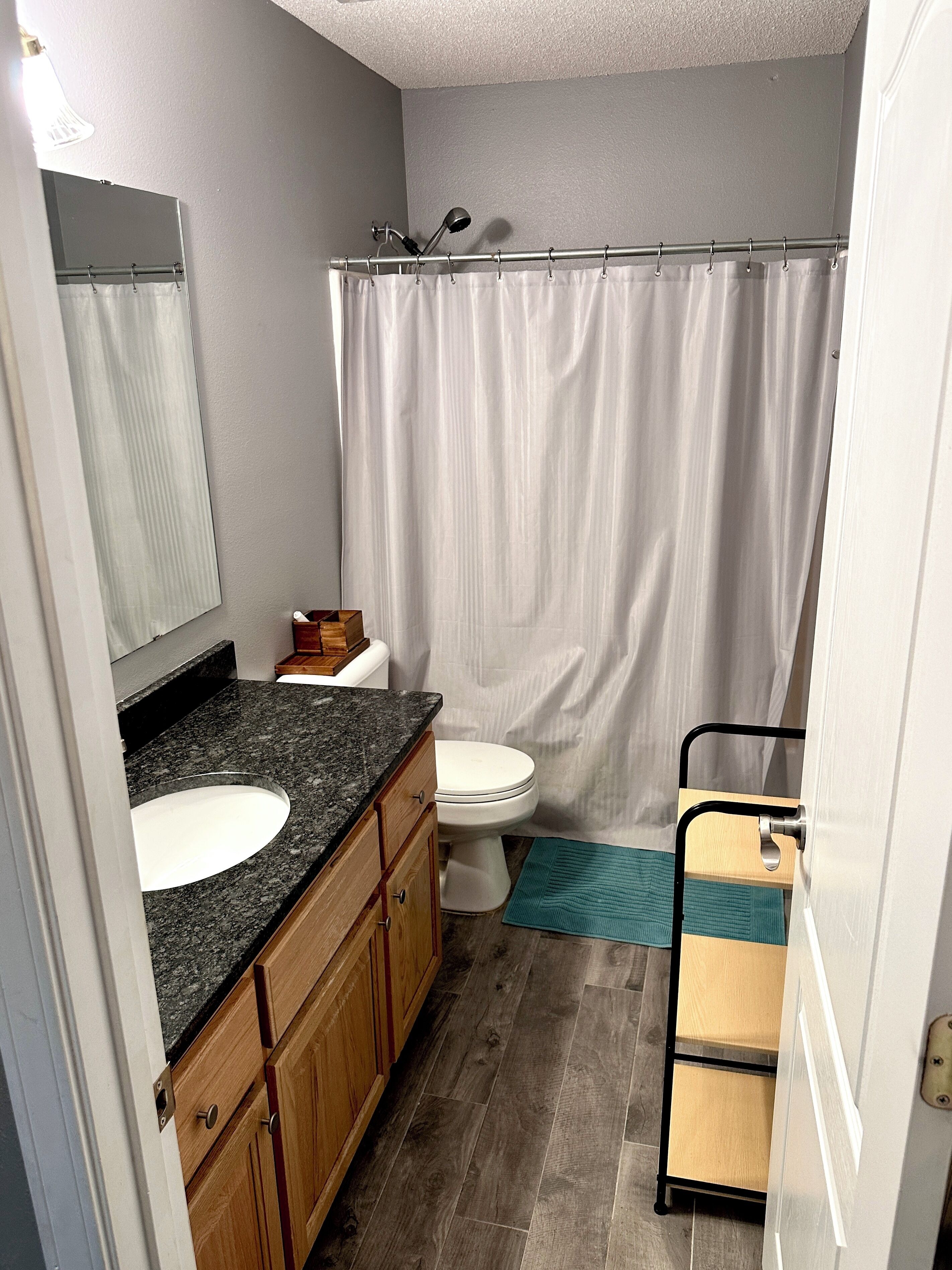 A bathroom with a white toilet, a sink with a granite countertop, wooden cabinets, a shower curtain, and a small rug.