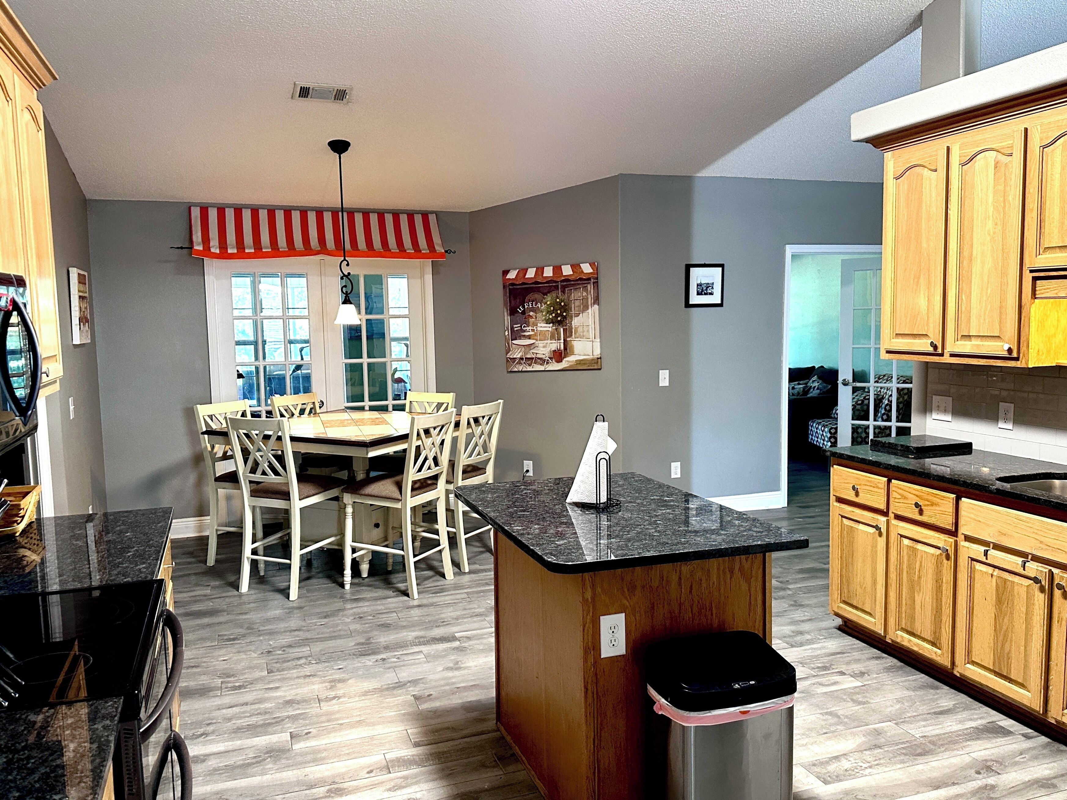 A kitchen with wooden cabinets, a granite countertop, a dining area with a table and chairs, and a window with a red and white striped valance.
