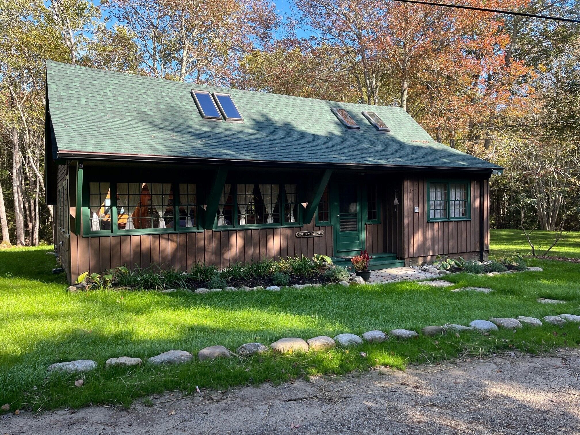 A rustic cabin with a green roof, two windows with shutters, and a porch with a sign reading "Stillmeadow".
