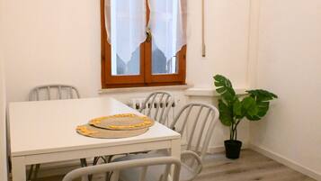 A dining area with a white table, chairs, a potted plant, and a window with curtains.