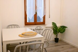 A dining area with a white table, chairs, a potted plant, and a window with curtains.