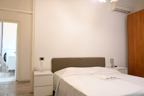 A bedroom with a bed, bedside table, nightstand, and an air conditioning unit.