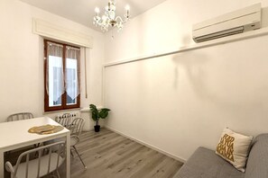 A small, well-lit room with a dining table, chairs, a potted plant, a sofa, and a chandelier.