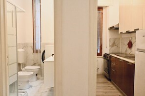 A narrow hallway leading to a bathroom with two toilets and a sink, a kitchen with a stove and cabinets, and a marble floor.