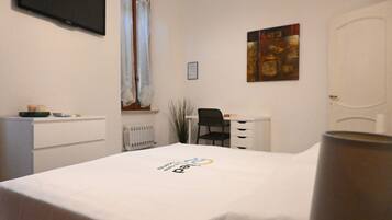 A hotel room with a bed, a desk, a chair, a radiator, a television, and a painting on the wall.