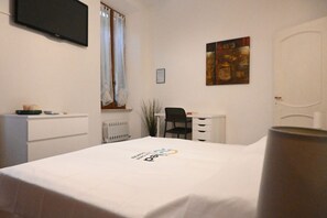 A hotel room with a bed, a desk, a chair, a radiator, a television, and a painting on the wall.