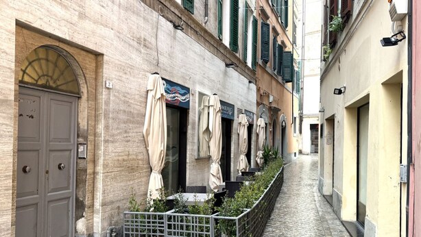 A narrow cobblestone alley with a row of outdoor seating areas, a metal gate, and a building with a large door and a number sign. - A42 - Ancona, two-room apartment in the center (Ancona)