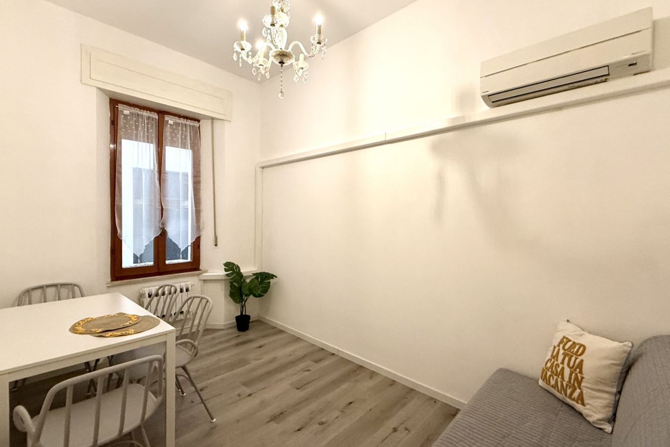 A small, well-lit room with a dining table, chairs, a potted plant, a sofa, and a chandelier.