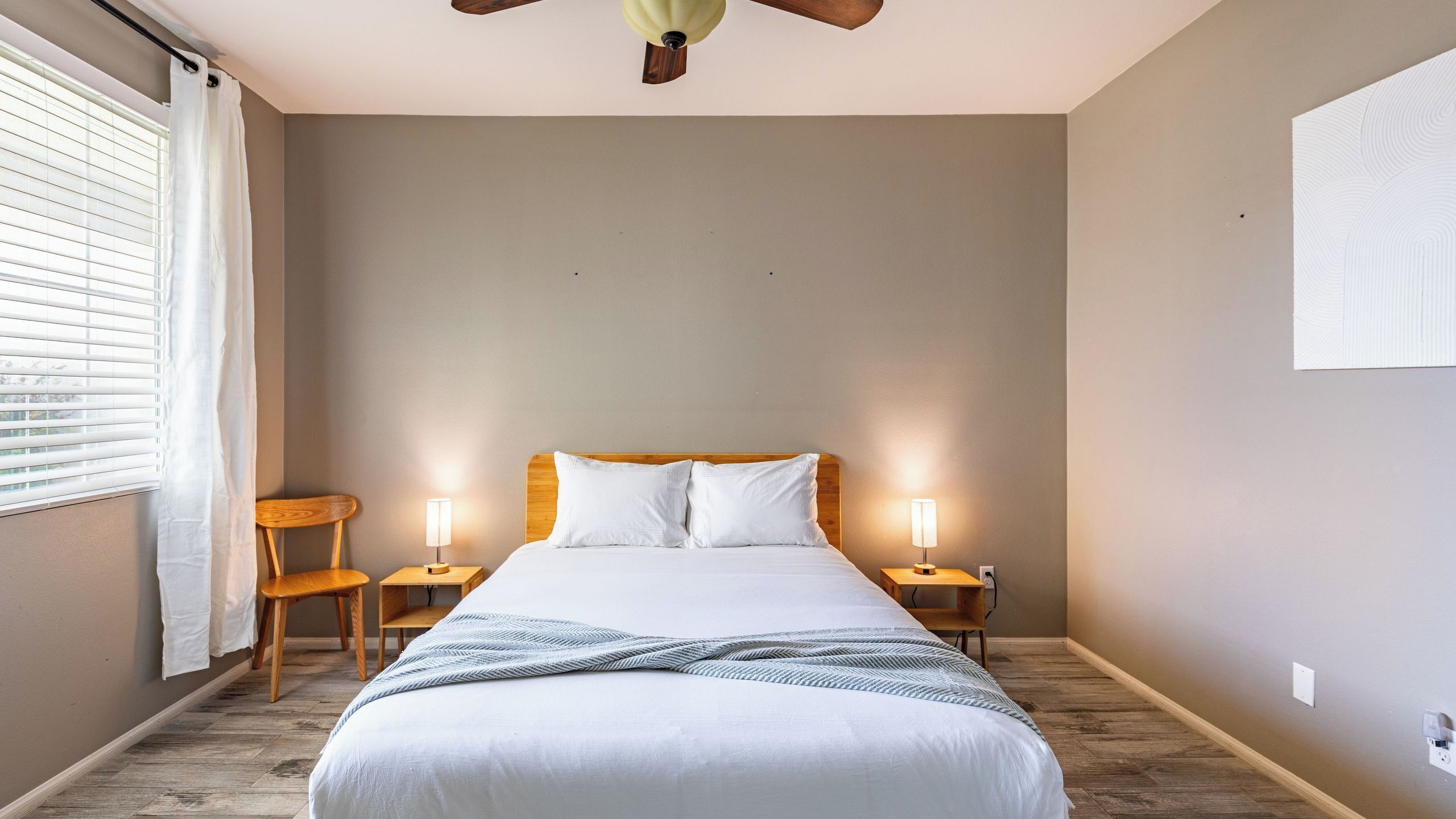 A bedroom with a bed, two bedside tables with lamps, a wooden chair, a ceiling fan, and a window with blinds.