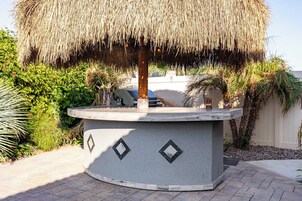 A circular stone structure with a thatched roof, surrounded by greenery and a white wall.