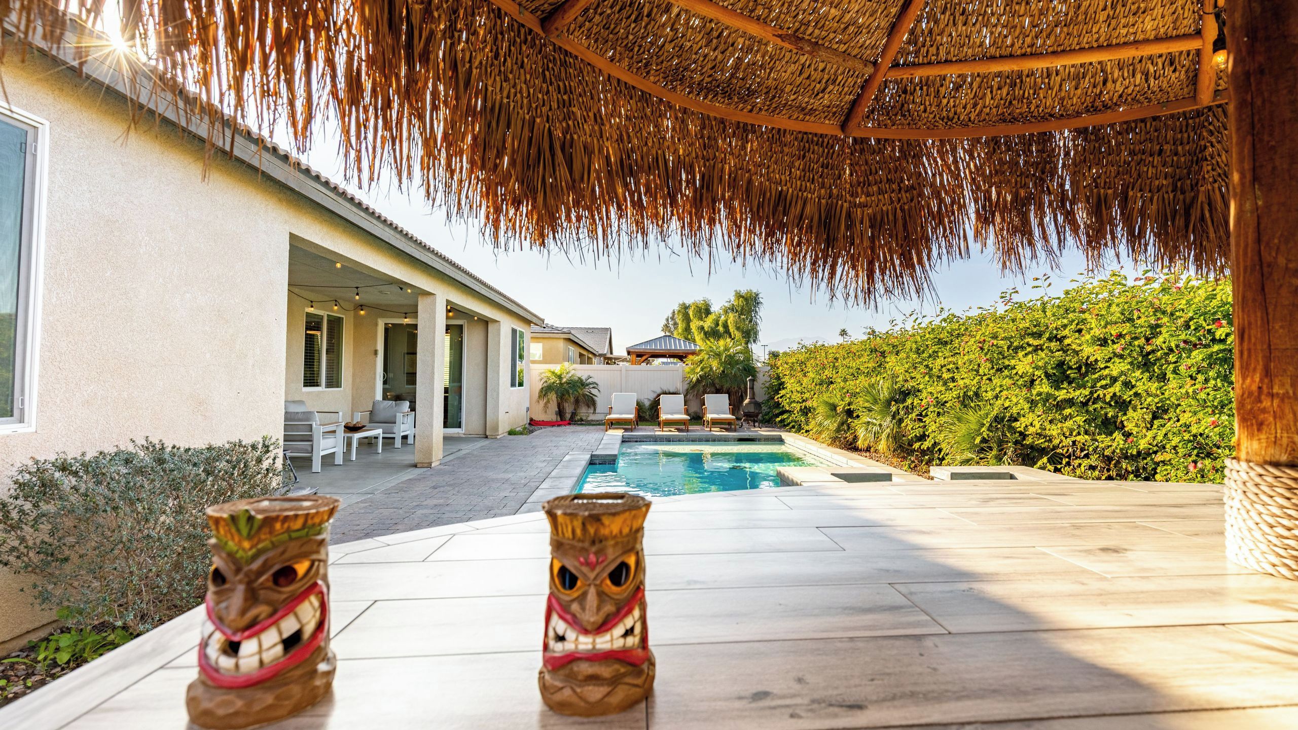 A pool area with tiki statues, a covered patio, and a view of a house with outdoor seating.