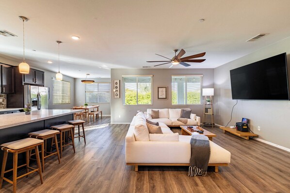 A modern living room with a large sofa, a dining area with stools, a kitchen with a bar, and a flat-screen TV mounted on the wall. - Luxe Indio Stay w/ Pool, Tiki Bar & Fire Pit Near Coachella Longfellow (Indio)