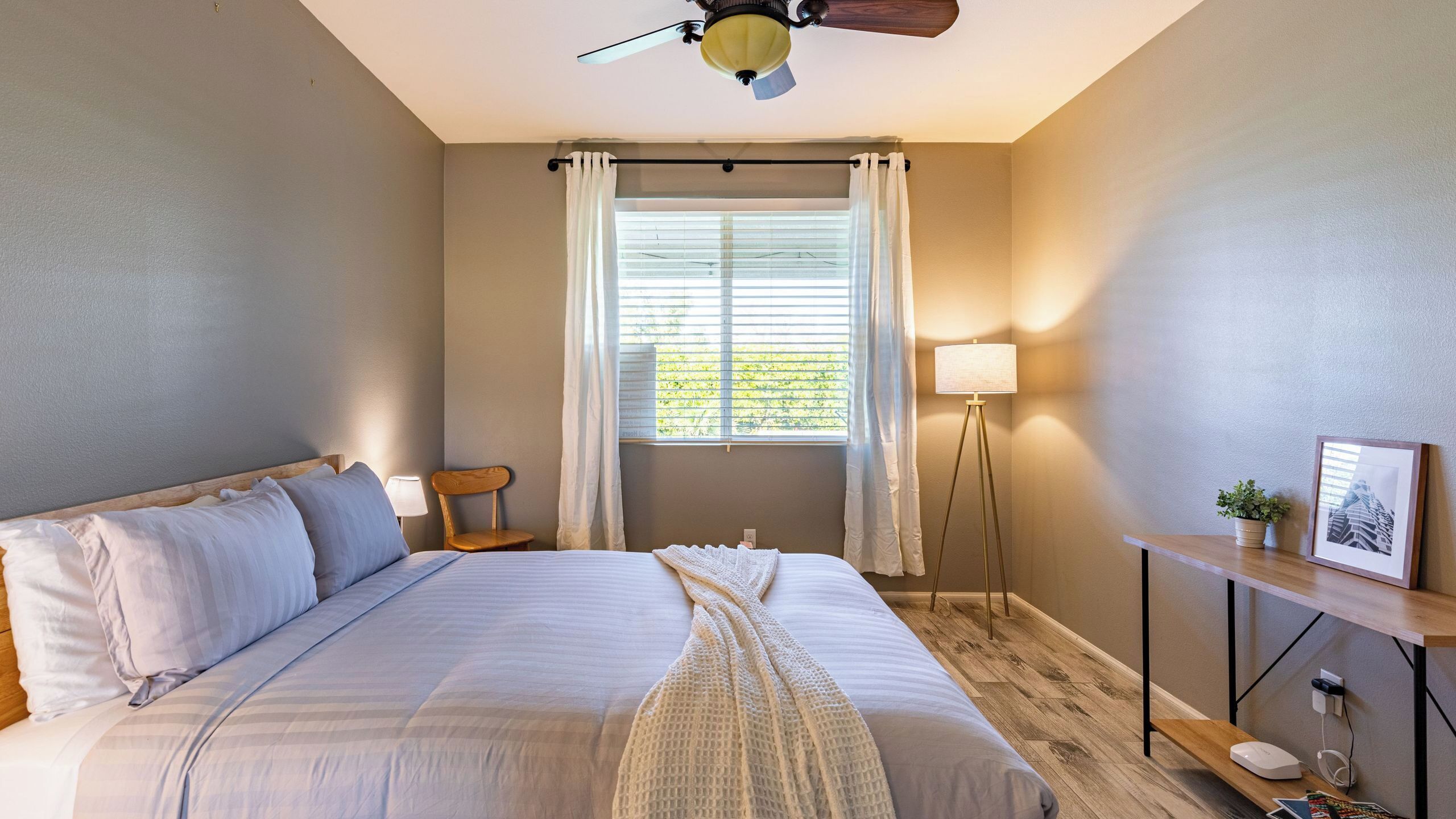 A bedroom with a bed, a chair, a floor lamp, a bedside table, and a window with curtains.