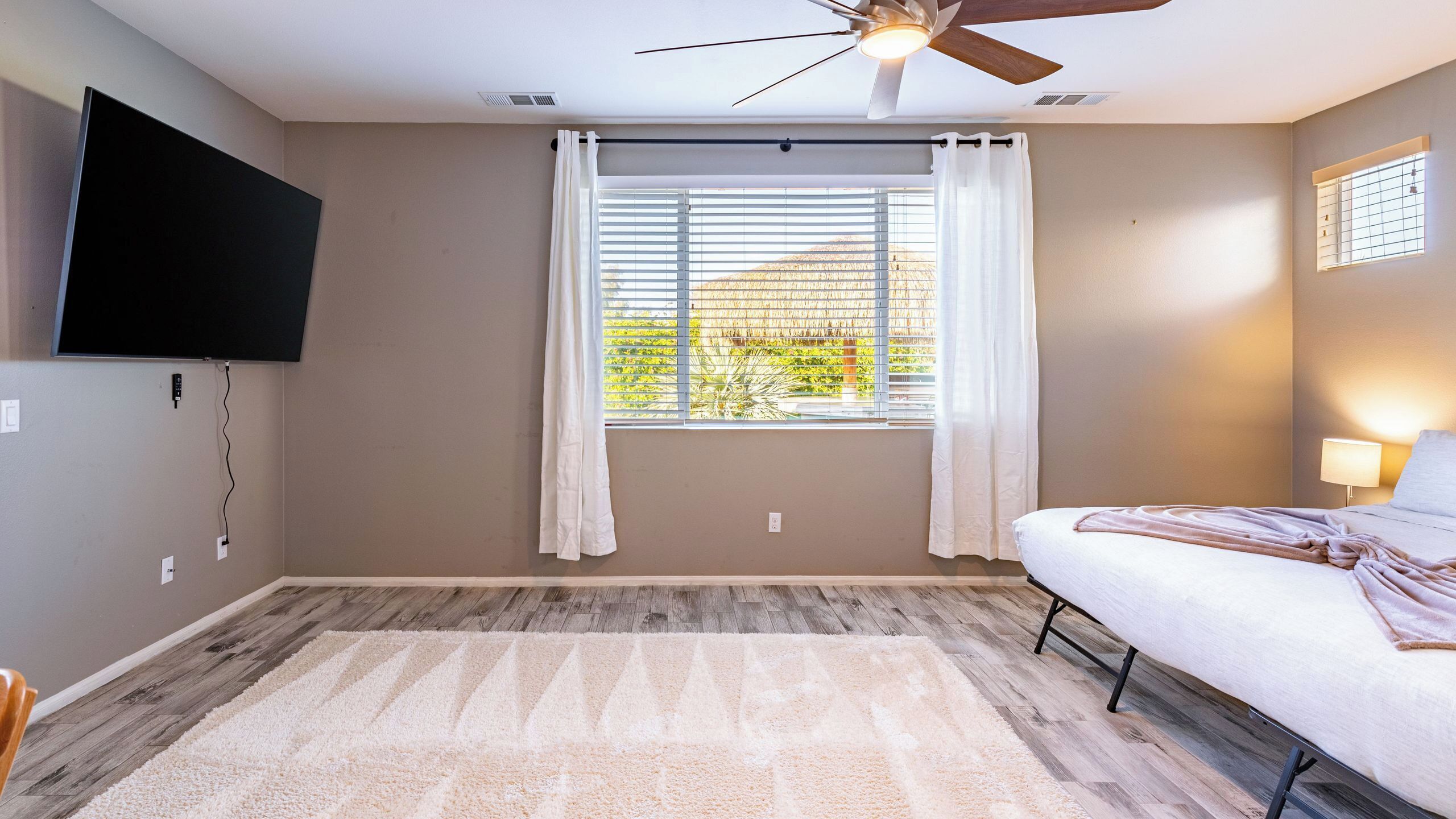 A bedroom with a ceiling fan, a large window with blinds, a TV mounted on the wall, and a bed with a blanket.