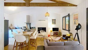 A spacious living area with a grey sofa, wooden dining table, and chairs. A kitchen is visible in the background with white cabinets and a large window.
