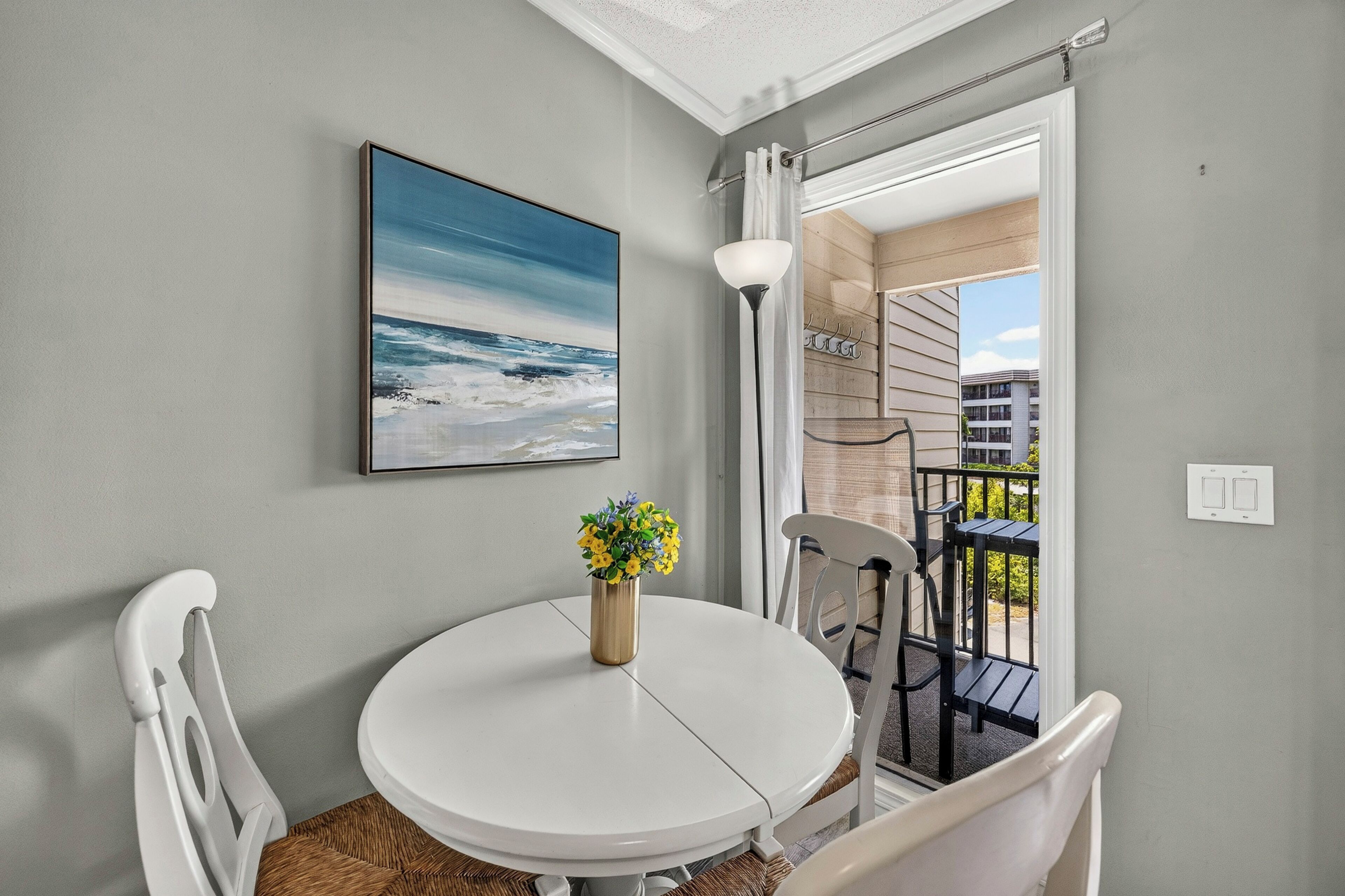 A room with a round table, white chairs, a painting of the ocean, a vase with flowers, and a view of a balcony with outdoor furniture.