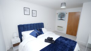 A neatly made bed with a blue headboard, white bedding, and a blue throw blanket. There are two bedside tables with lamps and a small drawer. The room has white walls, a wooden door, and a painting on the wall. - Luxury 2 Bedroom Apartment in Birmingham Next to Utility Arena (Birmingham)
