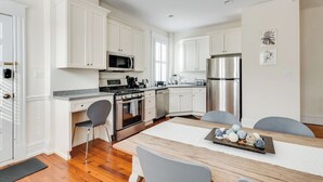 Una cocina moderna con armarios blancos, electrodomésticos de acero inoxidable y un comedor con mesa y sillas de madera.