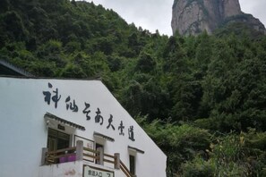 Un bâtiment orné d’une enseigne en caractères chinois, entouré d’une végétation luxuriante et dont l’arrière-plan est dominé par une formation rocheuse imposante.