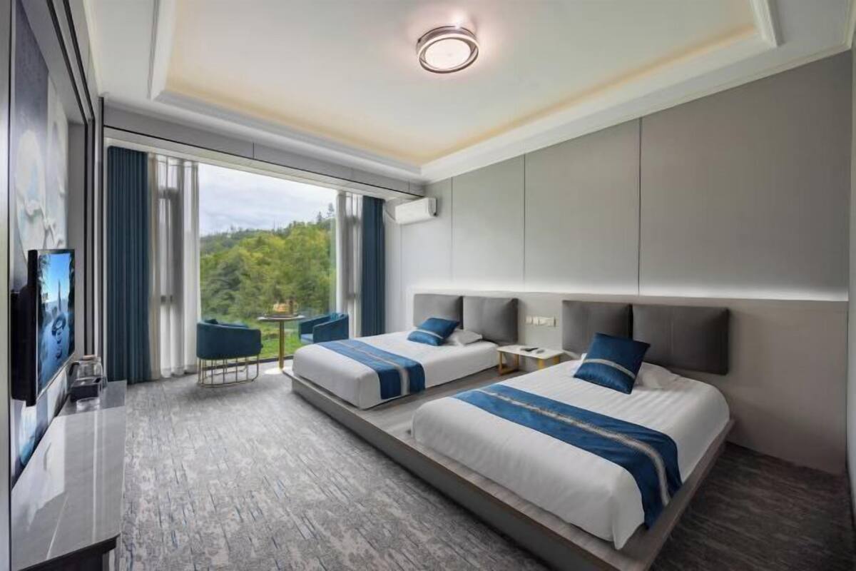 Habitación de hotel con dos camas, televisión, escritorio y balcón con vistas a zonas verdes.