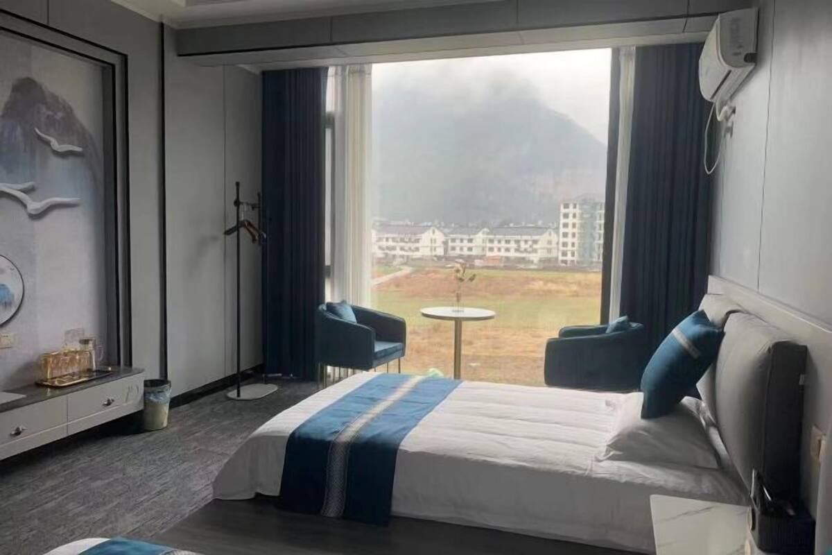 Habitación de hotel con cama, dos sillas, una mesita y vistas a edificios y montañas.