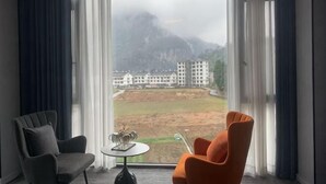 A room with a large window, two chairs, a small table, and a view of buildings and mountains.