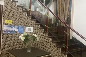 Una escalera con barandilla, una pared con azulejos y un código QR, y un jarrón con flores sobre una mesa.