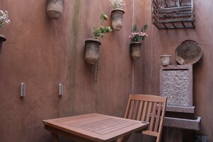 Un conjunto de mesa y sillas de madera frente a una pared con textura y macetas colgantes con flores.