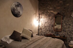 Un dormitorio con pared de piedra, un cuadro de luna redonda, un espejo y una mesita de noche con libros y una lámpara.