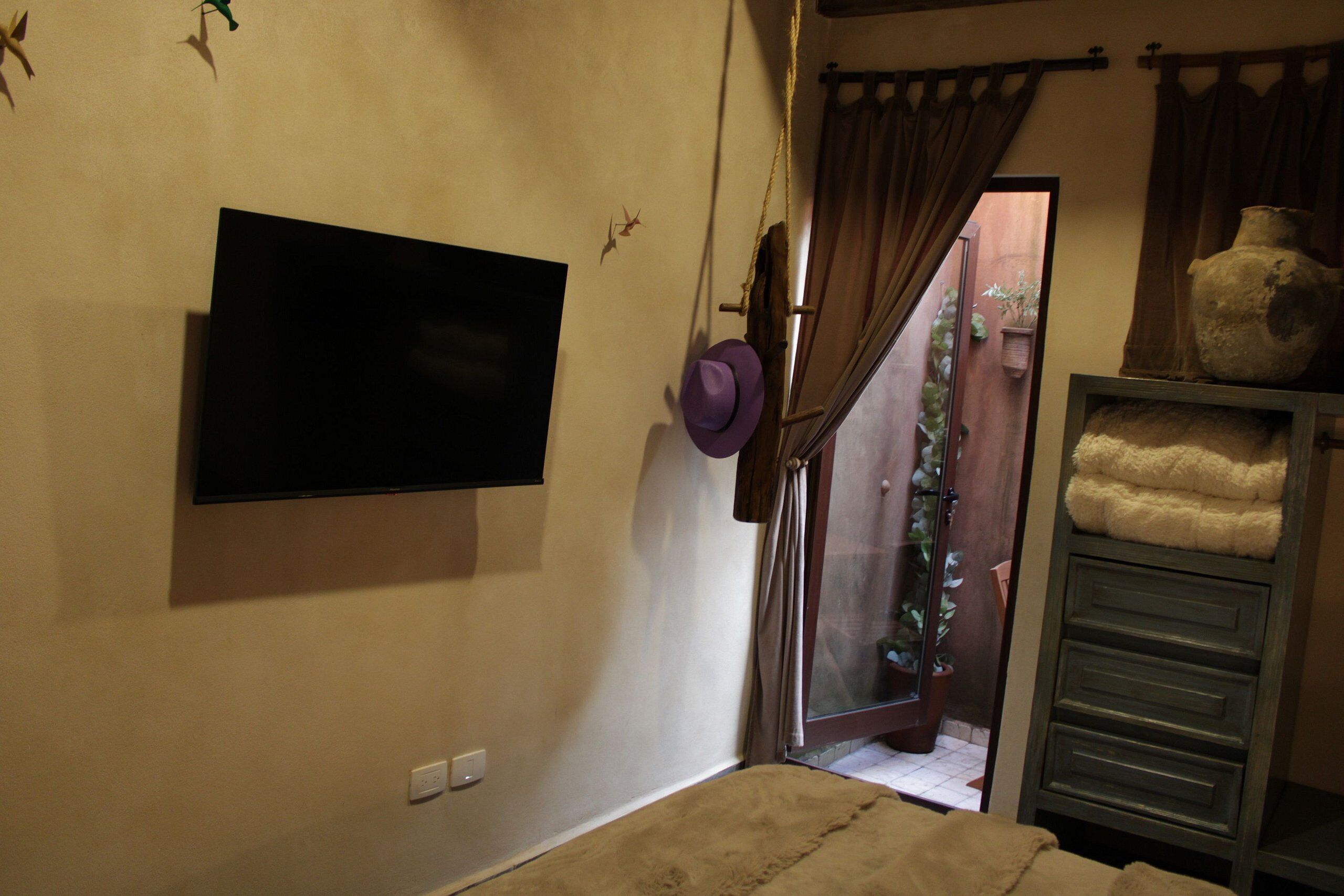 Una habitación con una televisión de pantalla plana montada, una cama con una manta color beige, un armario de madera y una planta en maceta cerca de la entrada.