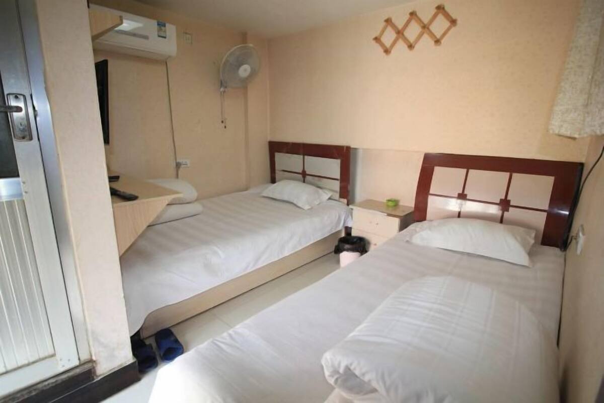 Une chambre d’hôtel avec deux lits simples, une table de chevet avec une lampe, un ventilateur fixé au mur et une petite table de nuit sur laquelle se trouve un objet vert.