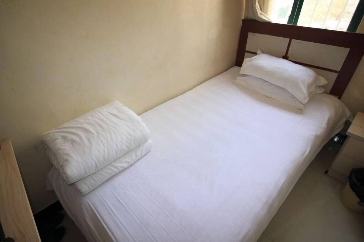 Un lit simple avec une literie blanche, dans une chambre dotée d’une tête de lit en bois et d’une fenêtre avec des rideaux.