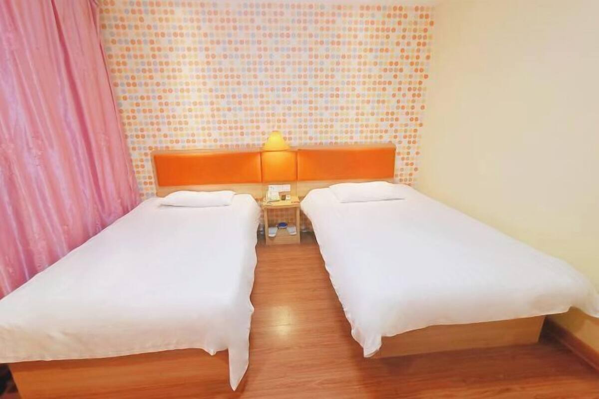 A room with two beds, a wooden floor, a patterned wall, and a pink curtain.