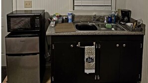 Una cocina compacta con microondas, refrigerador y lavamanos. Hay una ventana con persianas, una repisa con tazas y una toalla colgada del mueble.