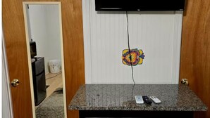 Una habitación pequeña con puerta de madera, un espejo, un televisor montado en la pared y una mesa con dos taburetes.