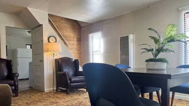 A living room with a patterned carpet, a blue armchair, a dining table with chairs, a potted plant, and a staircase.