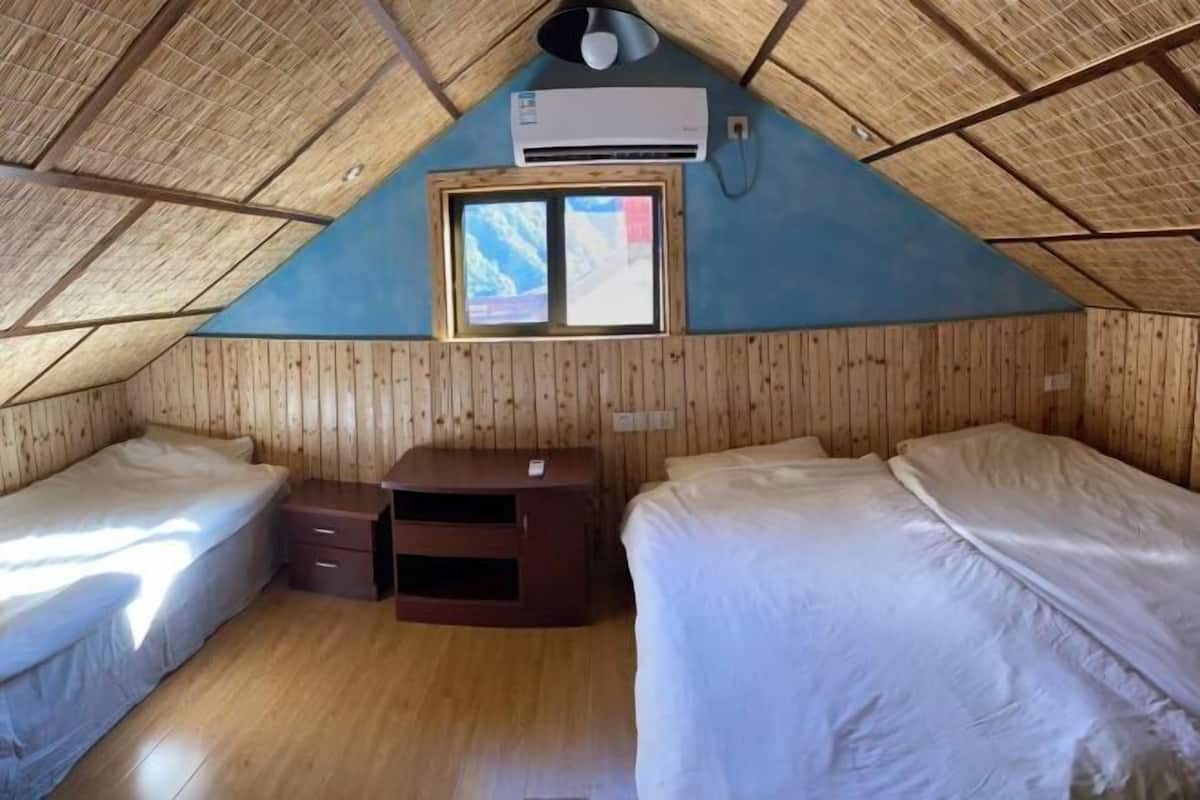 A triangular room with wooden walls and a sloped ceiling. There are two beds, a small desk, and an air conditioning unit mounted on the wall.