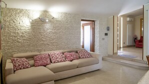 Un salon avec un mur en pierre, un canapé beige agrémenté de coussins à motifs, une table basse et, au fond, un fauteuil rouge.