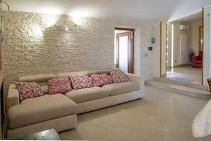 Ein Wohnzimmer mit Steinwand, einem beigefarbenen Sofa mit gemusterten Kissen, einem Couchtisch und einem roten Sessel im Hintergrund.