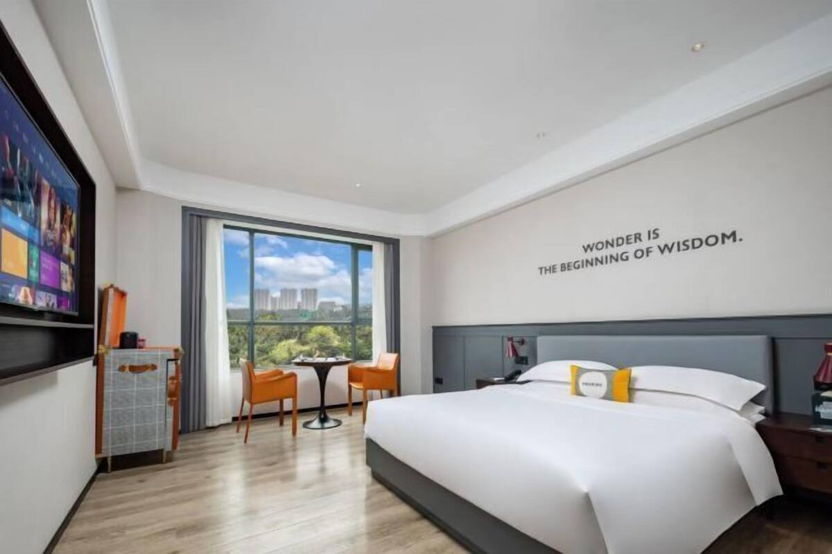 Una habitación de hotel moderna con una cama grande, un escritorio, una silla, un televisor y un ventanal con vista a la ciudad.