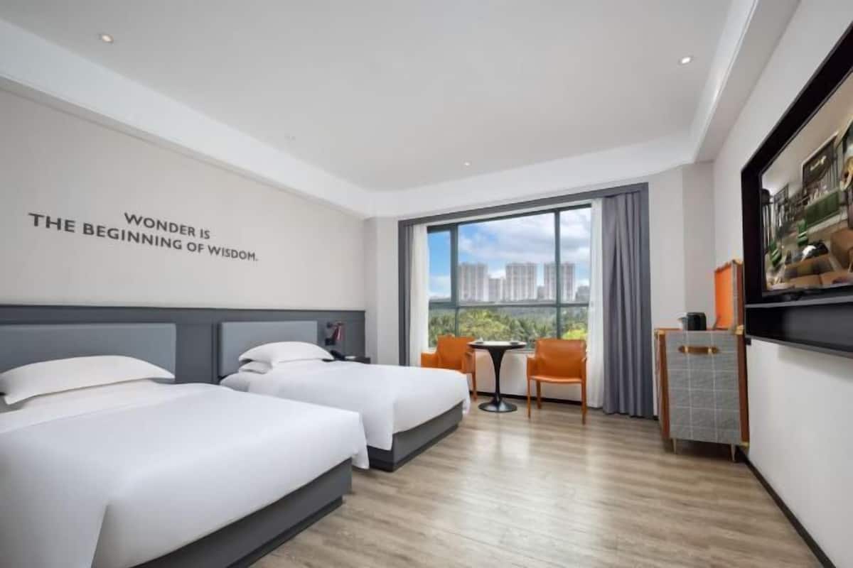 Habitación de hotel con dos camas, un escritorio, una silla, una mesita y un ventanal grande con vistas a edificios.